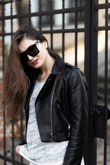Outdoor fashion portrait of stylish young woman having fun, emotional face , laughing, Urban city street style.Girl wearing white pants,t-shirt, black leather jacket , sunglasses and dark hat Fashion.