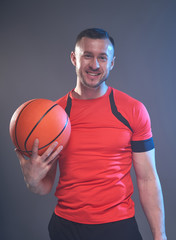 Young athletic man posing holding basketball ball with one hand in front of grey smoky background