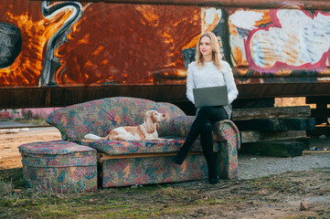Cheerful caucasian young freelancer girl sitting on old couch outdoor over rusty graffiti wall on...