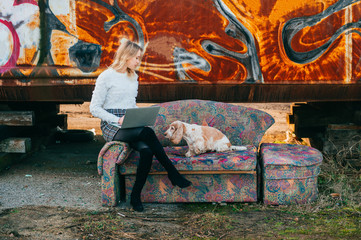 Obraz na płótnie Canvas Cheerful caucasian young freelancer girl sitting on old couch outdoor over rusty graffiti wall on background and using on laptop with her lovely cocker spaniel dog lying close to her.
