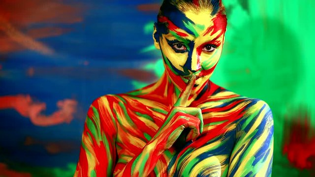 Color face art and body paint on woman holding her finger to her lips in a gesture for silence.. Abstract portrait of the bright beautiful girl with colorful make-up and bodyart.