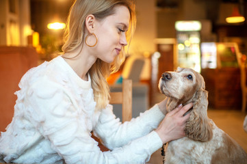 Young blonde girl in white sweater caring her lovely cocker spaniel puppy at the table in caffe.