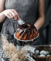 Woman hands holding chocolate Easter cake on white table decorated with spring flowers and wicker basket with eggs. Happy Easter holiday, selective focus