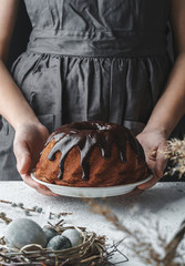 Woman hands holding chocolate Easter cake on white table decorated with spring flowers and nest with eggs. Happy Easter holiday, selective focus, toning