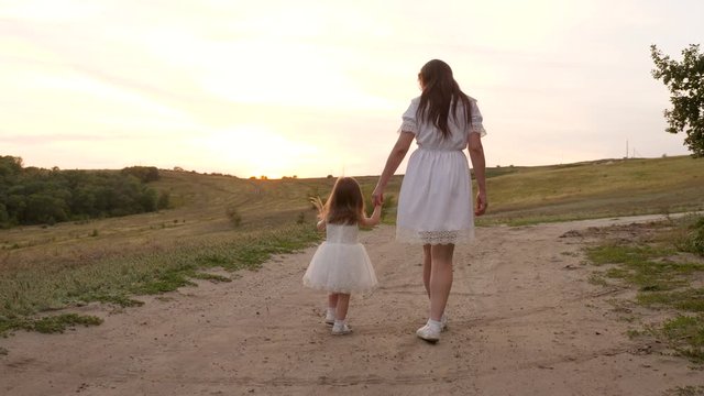 mom and child hold each other's hands and walk in the evening in the park. little daughter in a white dress is walking with her mom's hand along the road. happy family travels out of town