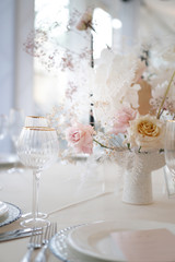Decor of a wedding luxury dinner in white and pink colors. Served tables with silver and gold objects, plates, glasses and cutlery. Fresh flowers roses in vases.