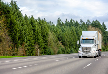 Fototapeta na wymiar White big rig semi truck with grille guard transporting cargo in refrigerator container running on the interstate wide highway with green trees on the side