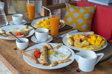 Tropical breakfast of fruit, coffee and scrambled eggs and banana pancake for two on the beach near sea. Top view, table setting.