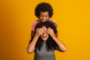 African American boy holding his mother closed eyes. Yellow background. Mothers Day. Brazilian...