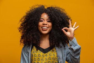 Beautiful curly-haired girl using her hands ok sign. Yellow background.