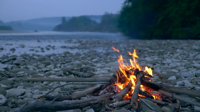 Close-up Shot Burning Firewood Campfire On Rocky River Shore. Evening Near Mountain Forest