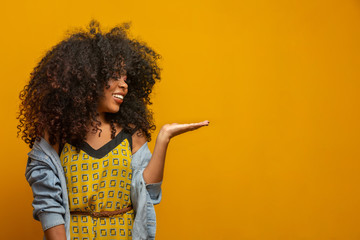 Cheerful Afro woman points away on copy space, discusses amazing promo, gives way or direction,...