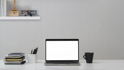 Close up view of modern workplace with blank screen laptop, office supplies and coffee cup on white table with shelf on white wall background