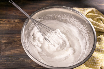 Fototapeta na wymiar Process of cooking meringue with whisk in metal bowl on wooden table. Recipe with whipped eggs, sugar. Perfect peaks for white sweet cookies, Italian or French dessert. Flat lay, copy space.