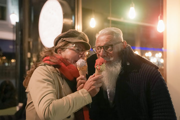 Elderly couple face to face eating ice cream in love attitude. Attractive retirees looking at each other having fun out of bar cafe gelateria. Concept of  romantic and love moment - Image