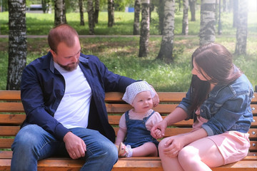 Family portrait mom and dad sitting on bench in city park with baby girl talking and smiling. Walking together at weekend. Mother, father, little girl spending time together. Baby care and parenthood.