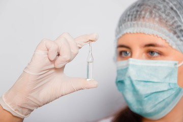 Close-up of a vaccine ampoule in the hands of a female doctor. Lab technician in a white coat, medical hat, mask and gloves, holds medicine in his hands.