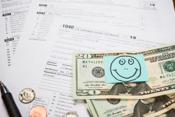 IRS 1040 tax return form with smiley face drawn on a sticker and attached to twenty dollar bill. Wage and tax statement concept