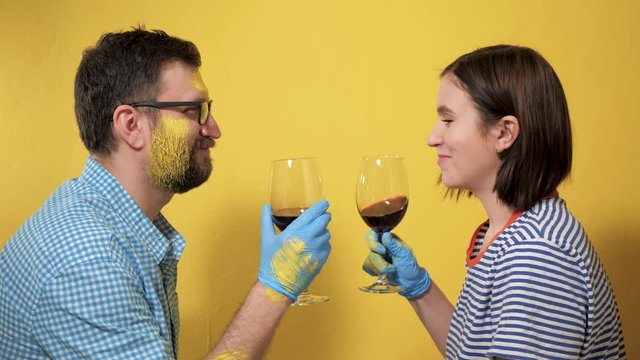 Man and girl sit opposite each other and solemnly drink wine. They are stained with yellow paint and very tired. Painting repair work over concept. Slow motion and close up