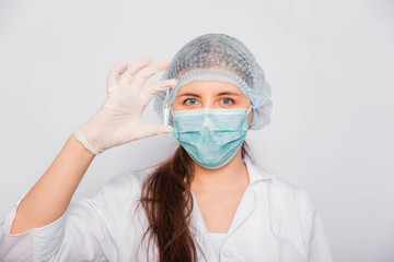  A female doctor in a white coat, medical cap, mask and gloves holds an ampoule with a vaccine.