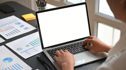 Behind shot through shoulder of smart man working as insurance statistic while typing on white blank screen laptop according information from graphic chart paper on working desk as background.