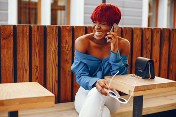 beautiful and stylish dark-skinned girl with red short hair, dressed in a blue top with long sleeves and white pants sitting in the cafe on the summer terrace at the table and using the phone