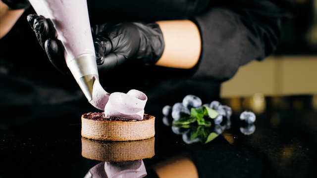 Pastry Chef Decorates Biscuit With Purple Cream From Pastry Bag, Close-up. Preparation Of Blueberry Cake At Commercial Bakery With Piping Bag