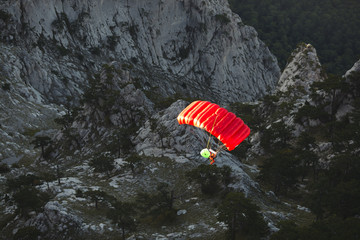 Base jumper under a bright red parachute canopy flies against a background of a dark rocky landscape after jumping from the top of a mountain, view from above.