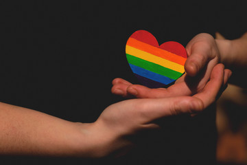 Female arms holding the heart coloured in LGBT pride colours on the dark background. Concept of the International Day Against Homophobia concept, sexual equality, feminism, social safety