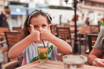 Happy little girl in a cafe on the street pb cocktail. The girl shows the figures with her hands. Walking and eating outside.