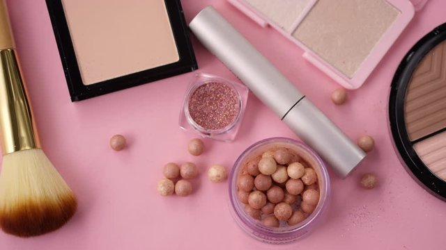 High angle shot of collection of makeup products on pink table. There is face palette, glitter, powder, highlighter, mascara, ball blusher and brushes