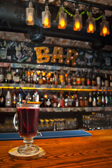 Red cocktail on bar counter. Drink with wine, fruit and cinnamon on the background of bottles with alcohol. Glass with booze on a wooden board. Cinnamon burns and smokes.