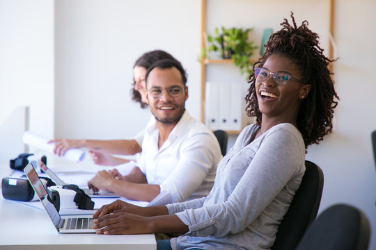 Cheerful colleagues laughing during work. Happy office workers sitting at table with VR headsets and laptops. Technology concept