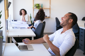 Laughing young man holding eyeglasses. Successful employee leaning back in chair with raised fist. Success, winning concept