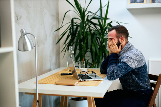 Young Caucasian tired bearded graphic designer sitting in his office and rubbing his eyes.
