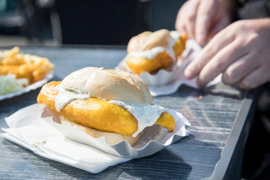 Mann isst Seelachs Backfisch im Bierteig als Fischbr&ouml;tchen mit Weizen Br&ouml;tchen und Remoulade drau&szlig;en auf Pappteller und Holztisch 