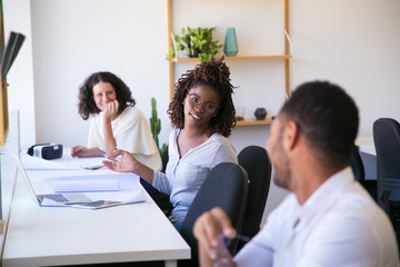 Fototapeta premium Smiling colleagues talking while sitting in office. Cheerful workers chatting at workplace. Communication and teamwork concept