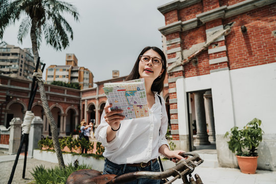 Portrait of asian chinese woman tourist hipster standing with map and bicycle in taichung city center in taiwan. Traveling summer vacation concept. young girl traveler in glasses reading paper guide - Powered by Adobe