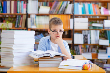 Cute boy with glasses in the library is reading a book. Piles of books are standing around.