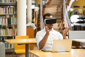 Obraz premium Focused guy with VR headset sitting in library. Serious African American man experiencing new VR glasses. Technology concept
