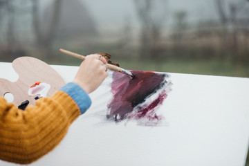 Close up photo of young female artist working on painting outdoors. She holds oil paints, artist brushes, canvas and palette. She is mixing colours on palette. Hands close up.