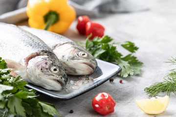 Two raw rainbow trout fish on a plate, greens and fresh vegetables for preparing healthy and tasty food. Healthy diet and delicious culinary concept. Copy space