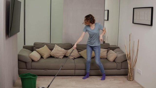 People, housework and housekeeping concept - happy woman in headphones with mop cleaning floor and dancing at home. Grey couch on the background. Slow motion
