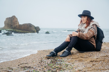 Beautiful red-haired woman in a hat and scarf with a backpack sits on the coast against the background of the rocks against the beautiful sea. Tourism, rest, lifestyle. Place for text or advertising