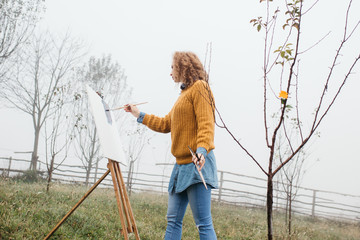 Young female artist working on painting outdoors. She is in front of the canvas and drawing.She holds oil paints, artist brushes, canvas and palette. Portrait of concentrated woman with curly hair. 