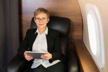 beautiful aged woman in the cabin of a private plane in a chair with a tablet in her hands