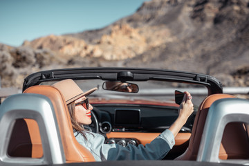 Young woman photographing on phone while traveling by convertible car on the picturesquare road on the desert valley