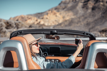 Young woman photographing on phone while traveling by convertible car on the picturesquare road on...