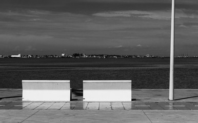 Black and white shot of the Luanda bay with buildings in the distance. Two benches. Noltalgic concept.