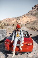 Lifestyle portrait of a carefree woman dressed casually in jeans and red hat sitting on the car trunk, enjoying road trip on the desert valley